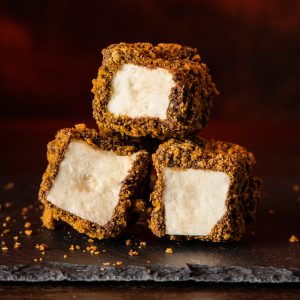 Three chocolate dipped square marshmallows rolled in graham cracker crumbs, one stacked on top of the two, on a gray slate surface.
