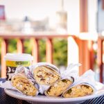 A plate of four Deli breakfast burritos wrapped in Deli paper with a cup of Zingerman's coffee behind it, and the Next Door Cafe front porch in the background.