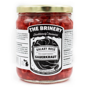 A jar of The Brinery Galaxy Rose Sauerkraut. The jar is wide and cylindrical with a gold lid and a black and white label. The sauerkraut inside is pink.