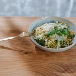 A blue ceramic bowl on a wooden countertop, filled with spaghetti topped with grated Parmagiano and fresh basil leaves, with a fork resting on the rim.