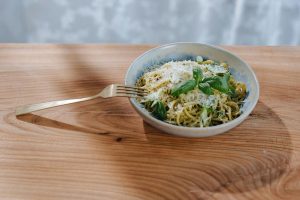 A blue ceramic bowl on a wooden countertop, filled with spaghetti topped with grated Parmagiano and fresh basil leaves, with a fork resting on the rim.