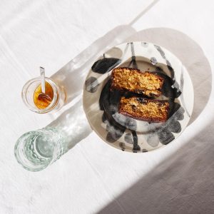 A top-down photo of two slices of fennel cake on a ceramic plate with a jar of preserves and a glass of water.