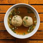 Consider the Matzo Ball A top-down photo of a bowl of two matzo balls in a bowl of broth topped with finely sliced green onions. The bowl is on a wooden table.