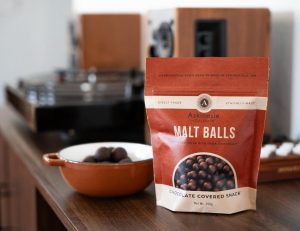A bag of Askinosie Malt Balls on a dark wooden countertop with a small ceramic bowl of malt balls behind it.