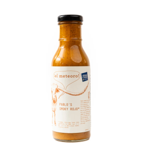 A photo of El Meteoro Smoky Rojo sauce. The bottle is clear and shows the red sauce inside. The label is parchment colored with text matching the color of the sauce, and an image of a cow with long horns and a speech bubble that says "El Meteoro!"