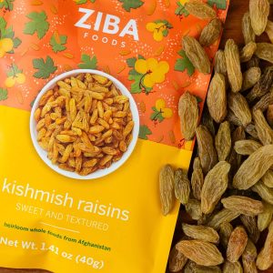 A close-up photo of a bag of Ziba Kishmish Raisins resting on a pile of Kishmish Raisins. The bag is solid yellow on the bottom half and orange with a floral pattern on the top half, and an image of a bowl of the raisins in the middle.